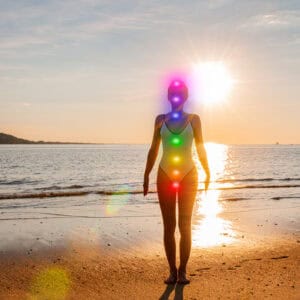 A woman pictured staning at waters edge at sunrise on a beach, in a one piece swimming costume, with different colours circles placed from her head to her pelvis, representing her 7 Chakra locations, something we work with in our energy healing at The Healing Practice